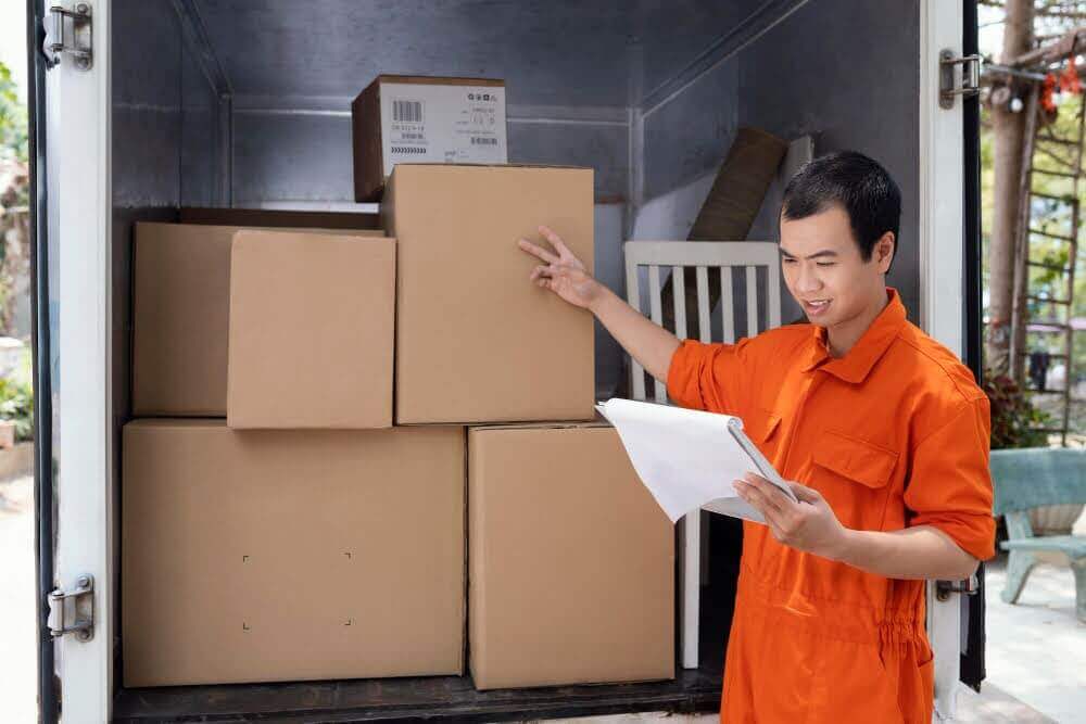 Delivery worker in an orange uniform verifying shipment details with a clipboard inside a loaded truck, illustrating the practical application of understanding logistics.