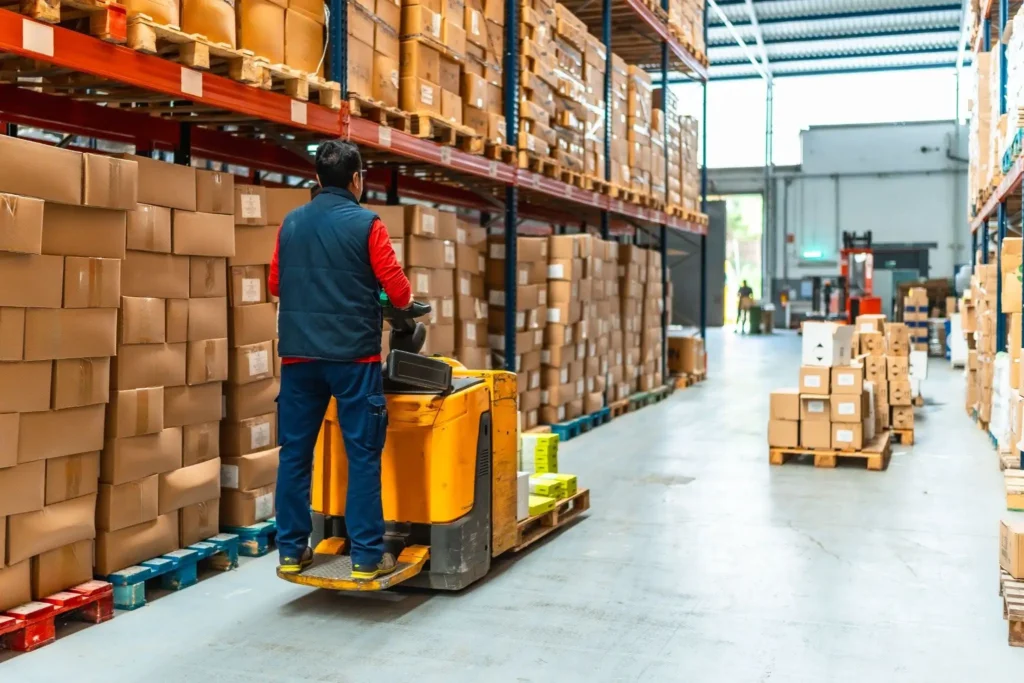 Warehouse worker on a yellow electric pallet jack moving boxes along a shelved storage aisle