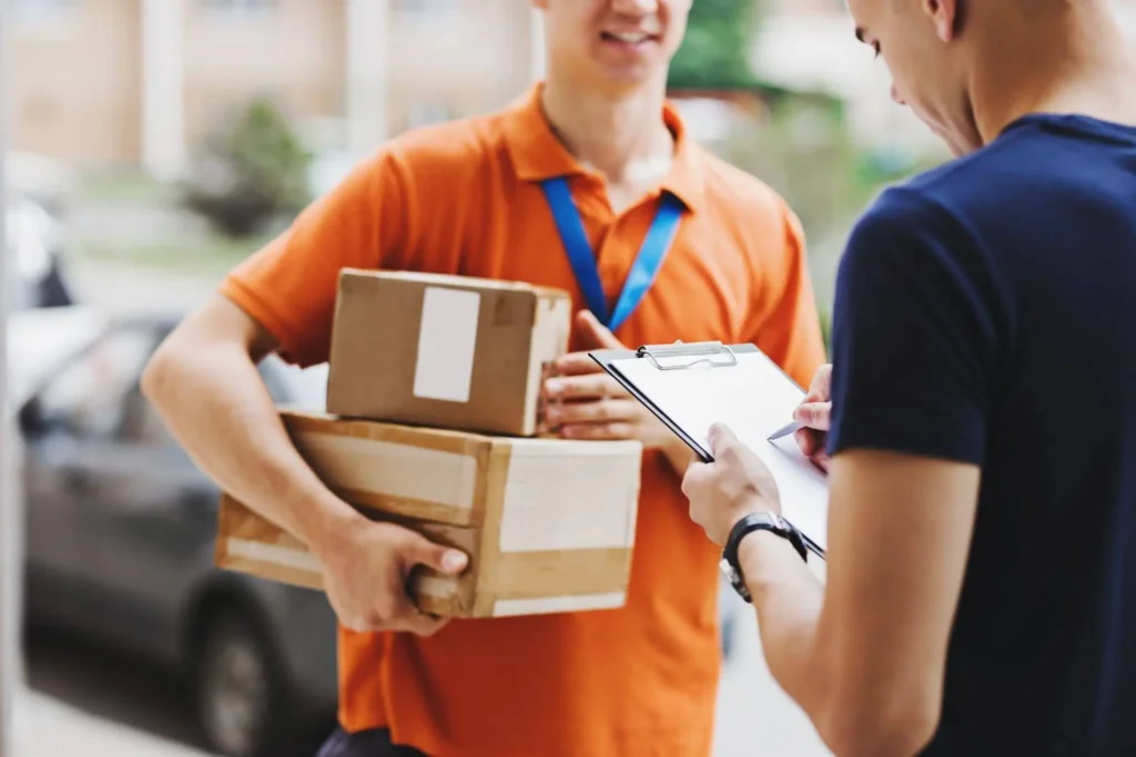 Delivery courier in an orange uniform handing parcels to a customer who signs a clipboard at the door.