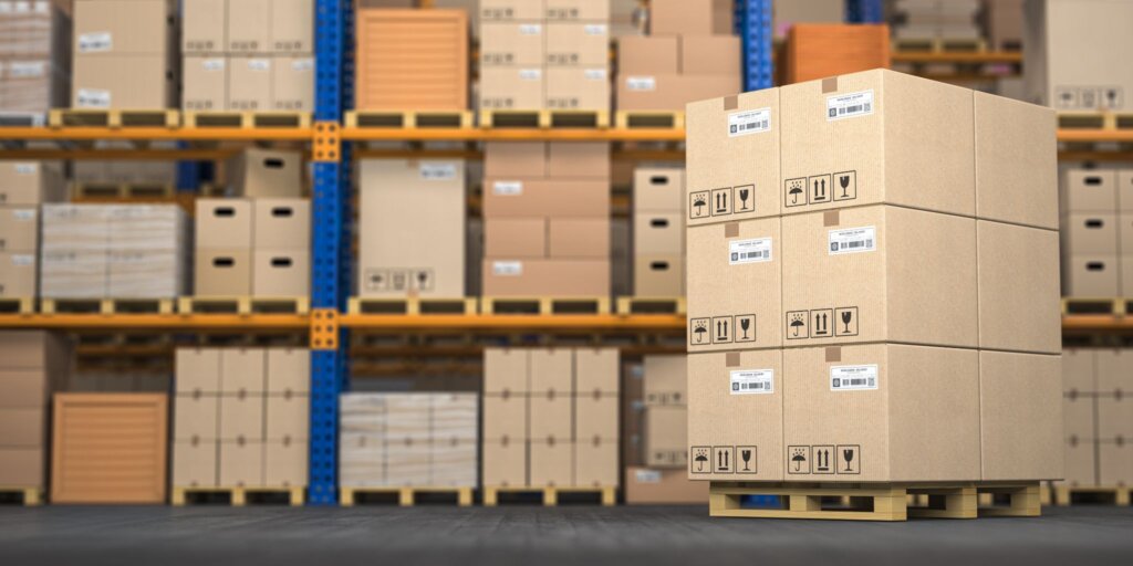 Stacked shipping boxes on a pallet with pallet racking shelves blurred in the background.