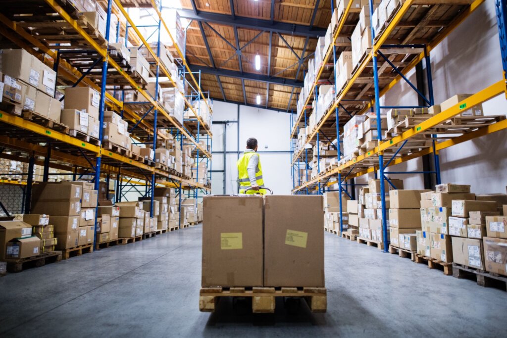 Warehouse worker pulling a loaded pallet jack down a tall aisle of pallet racking